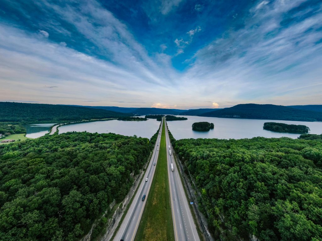 A long, straight highway cuts through dense green forest and spans a tranquil lake dotted with islands, under a dramatic, cloud-streaked sky.