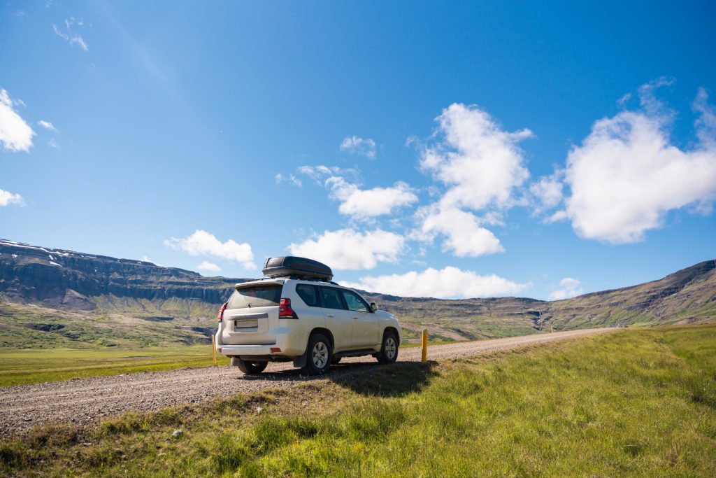 A white SUV with a rooftop cargo carrier is parked on a gravel road surrounded by vast green fields and rolling hills, with a clear blue sky and scattered white clouds overhead.