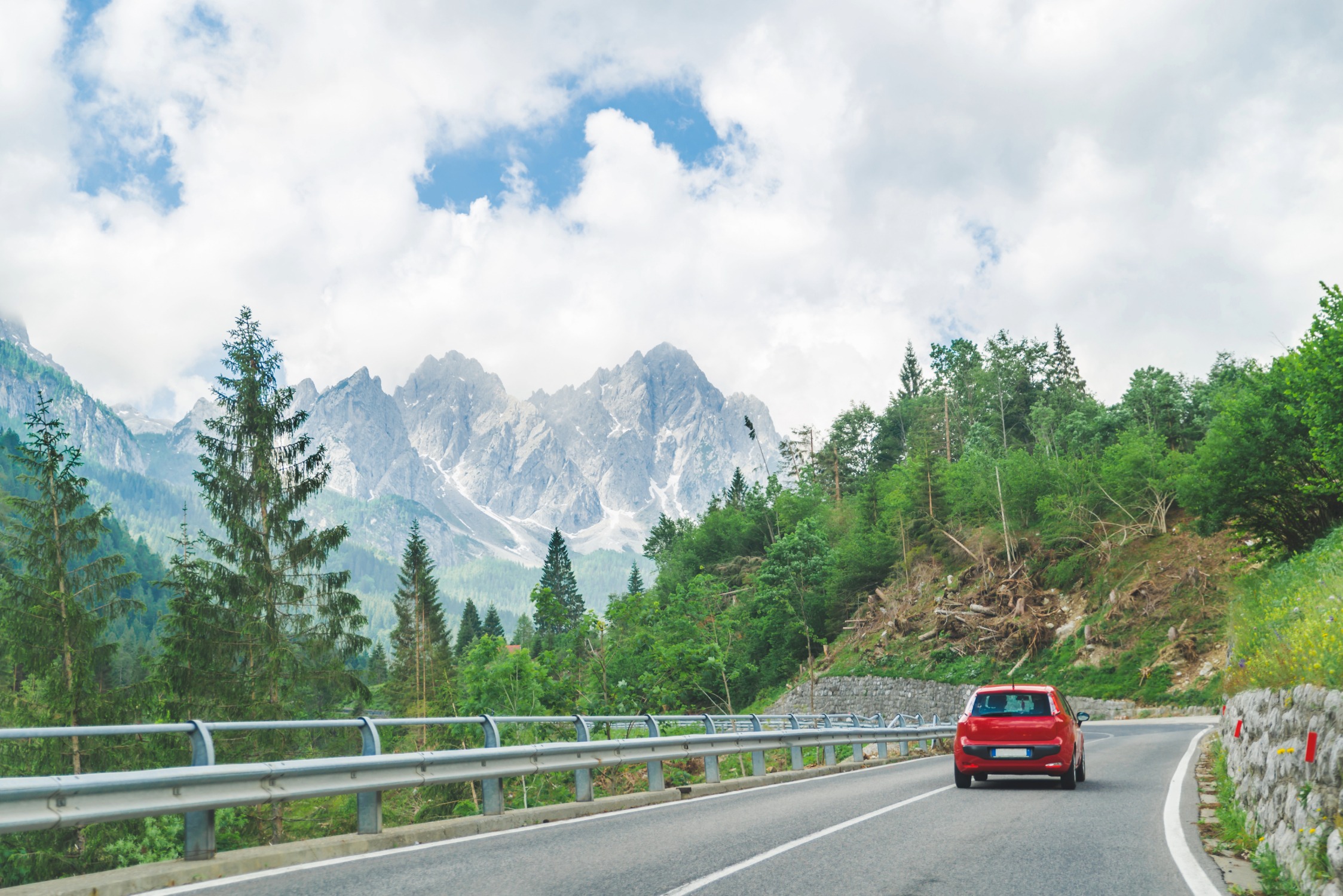 A red car drives along a winding mountain road bordered by guardrails and lush green trees, with towering, snow-dusted peaks rising dramatically in the background beneath a partly cloudy sky.