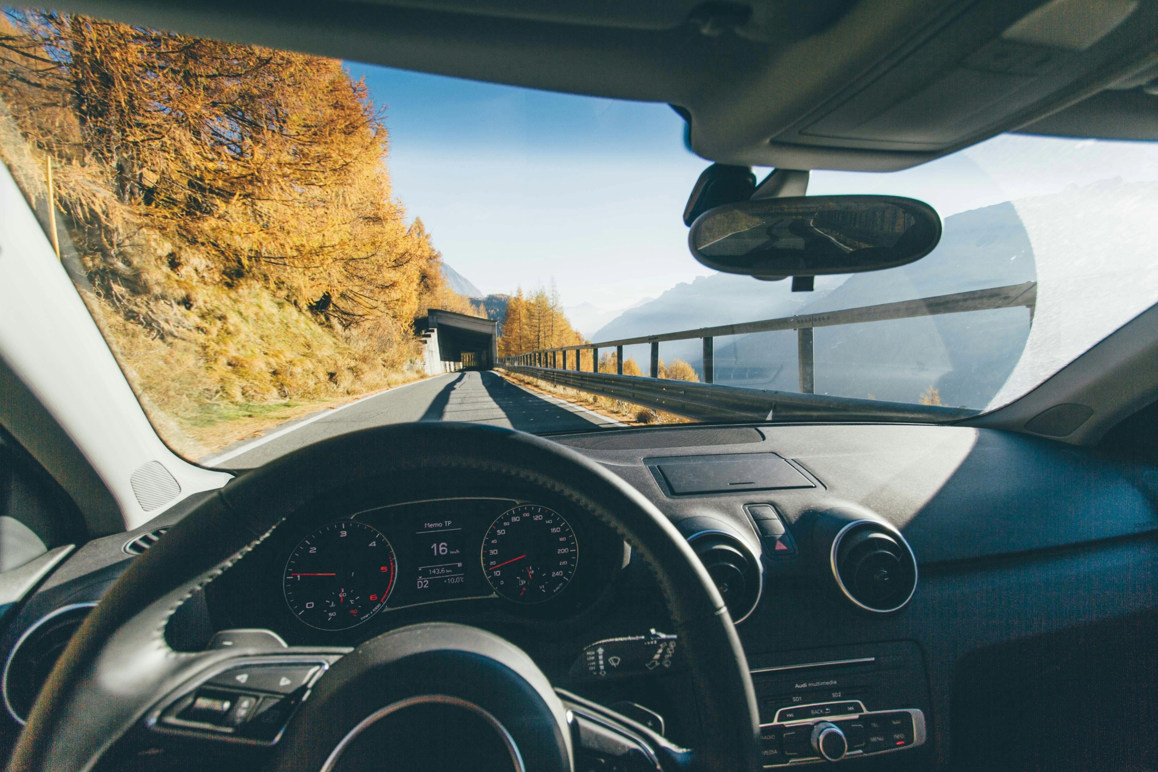 The image captures a scenic view from the driver’s seat of a car traveling cautiously along a winding mountain road, with the dashboard, steering wheel, and autumn landscape visible through the windshield.