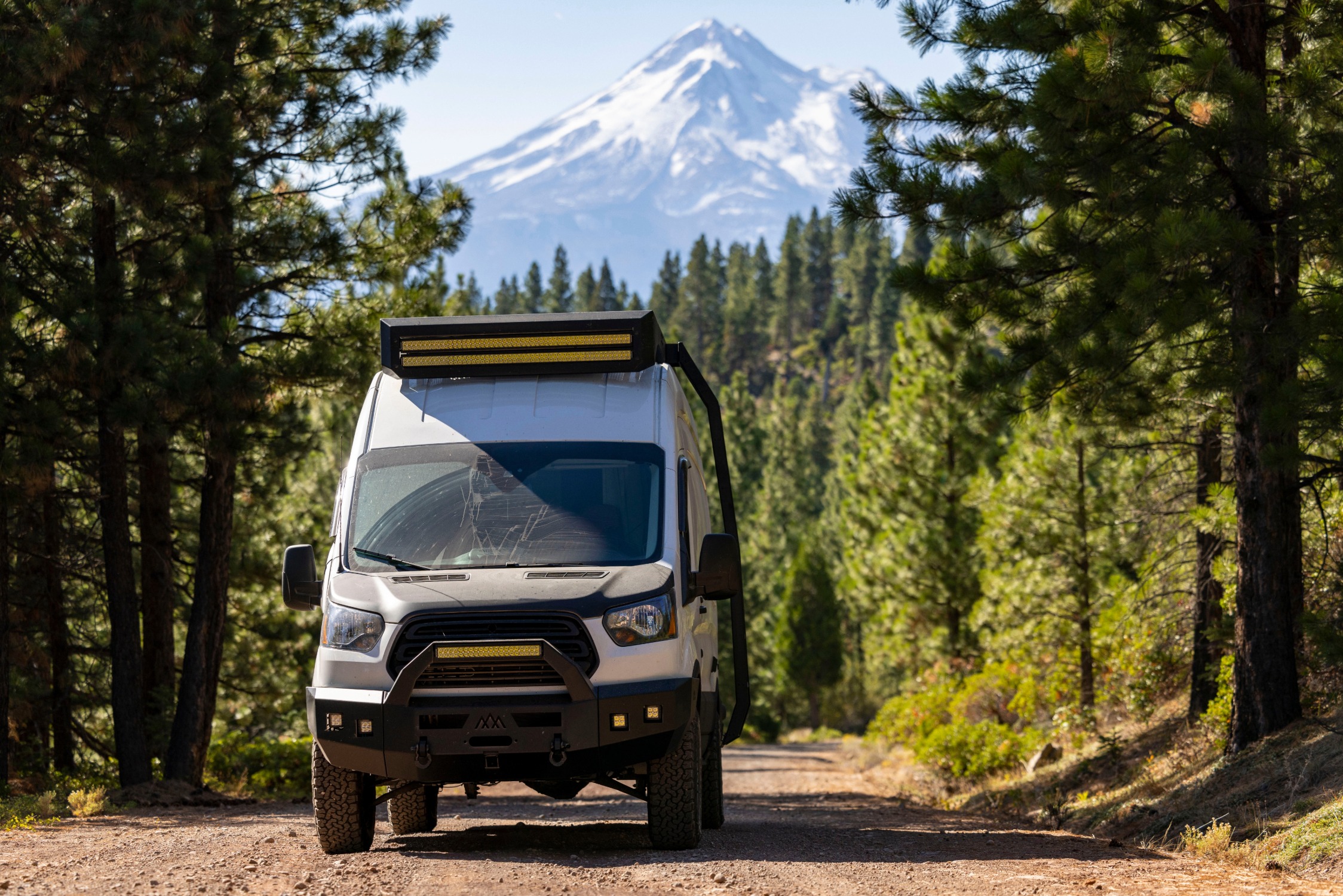 A white, modified Ford Transit van drives along a dirt road through a dense forest towards a snow-capped mountain in the distance.