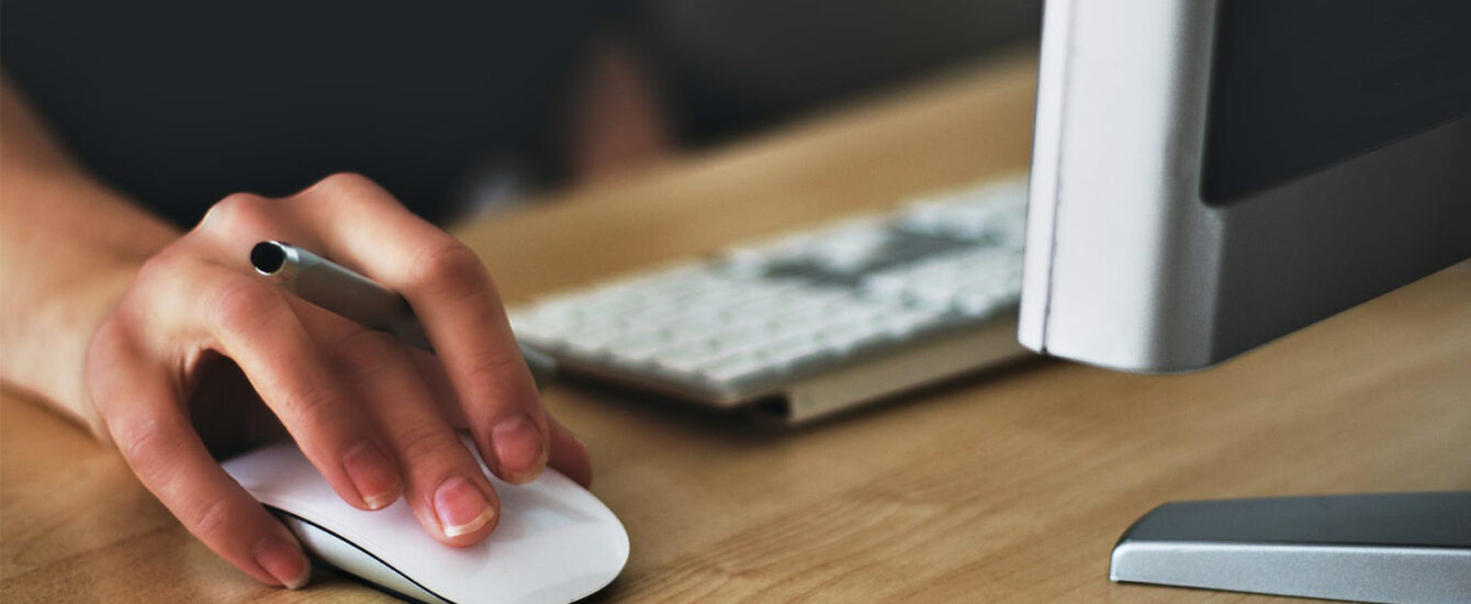 A person uses a white computer mouse while holding a pen, with a keyboard and monitor in view, suggesting a multitasking work environment.
