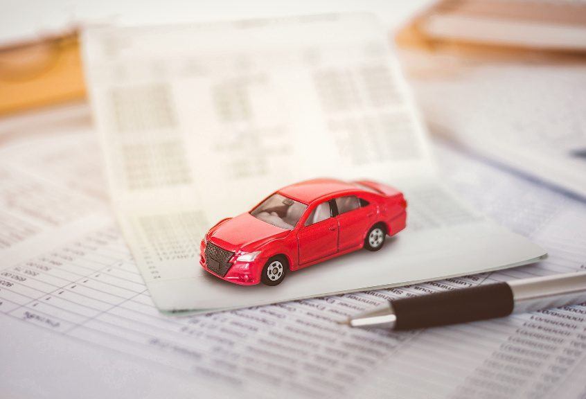 A small red toy car placed on top of a vehicle registration document, surrounded by financial papers and a pen. The image symbolizes car ownership, registration, and financial planning related to vehicles.