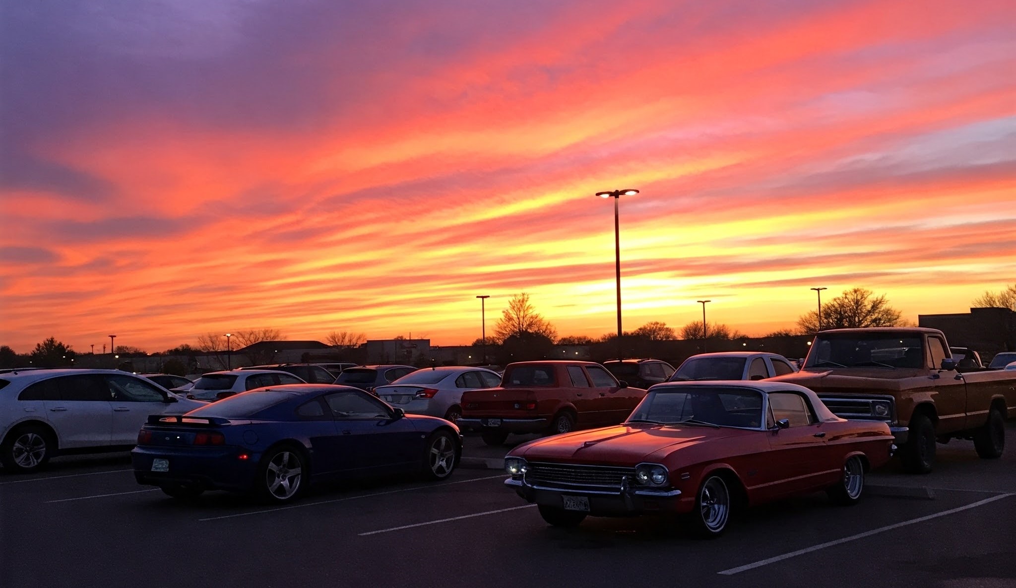 A vibrant sunset paints the sky in shades of orange pink and purple over a parking lot filled with cars.