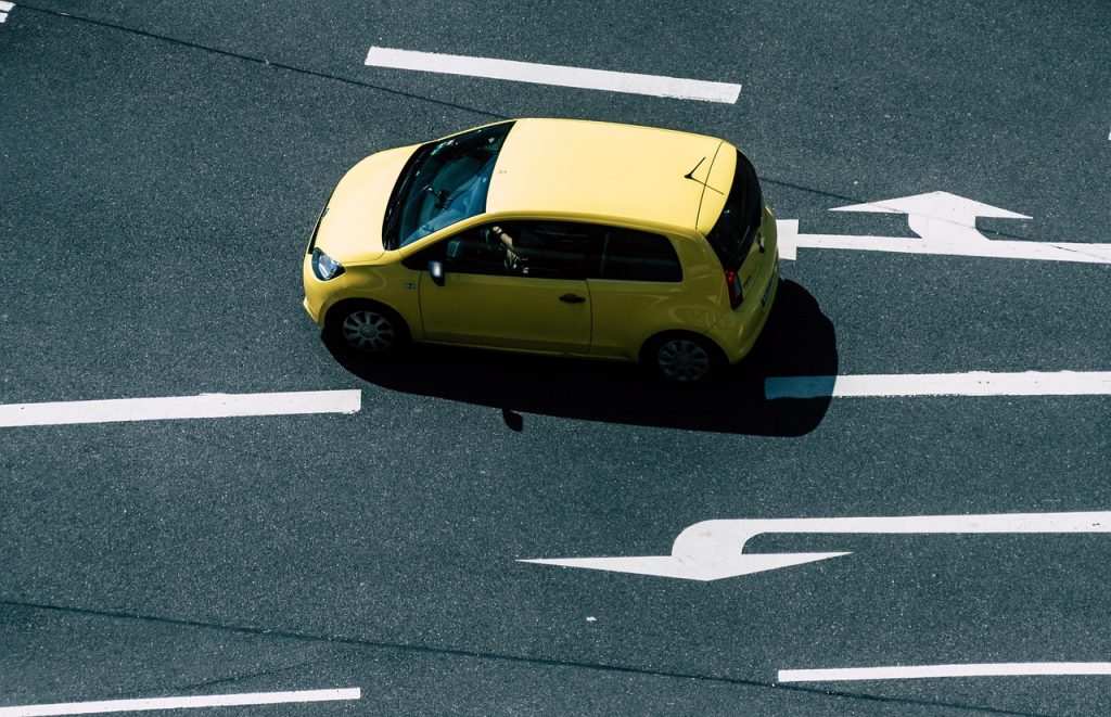 A small yellow car is driving alone on a multi-lane road marked with bold white directional arrows.