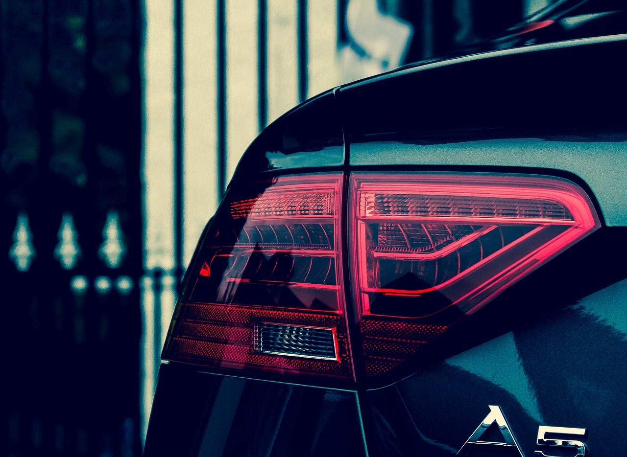 A close-up of a black Audi A5 shows its red taillight glowing against a background of metal bars.