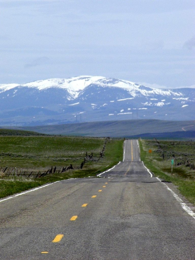 A long, open highway stretches through Montana's vast plains toward snow-capped mountains, capturing the state's rugged and scenic beauty.