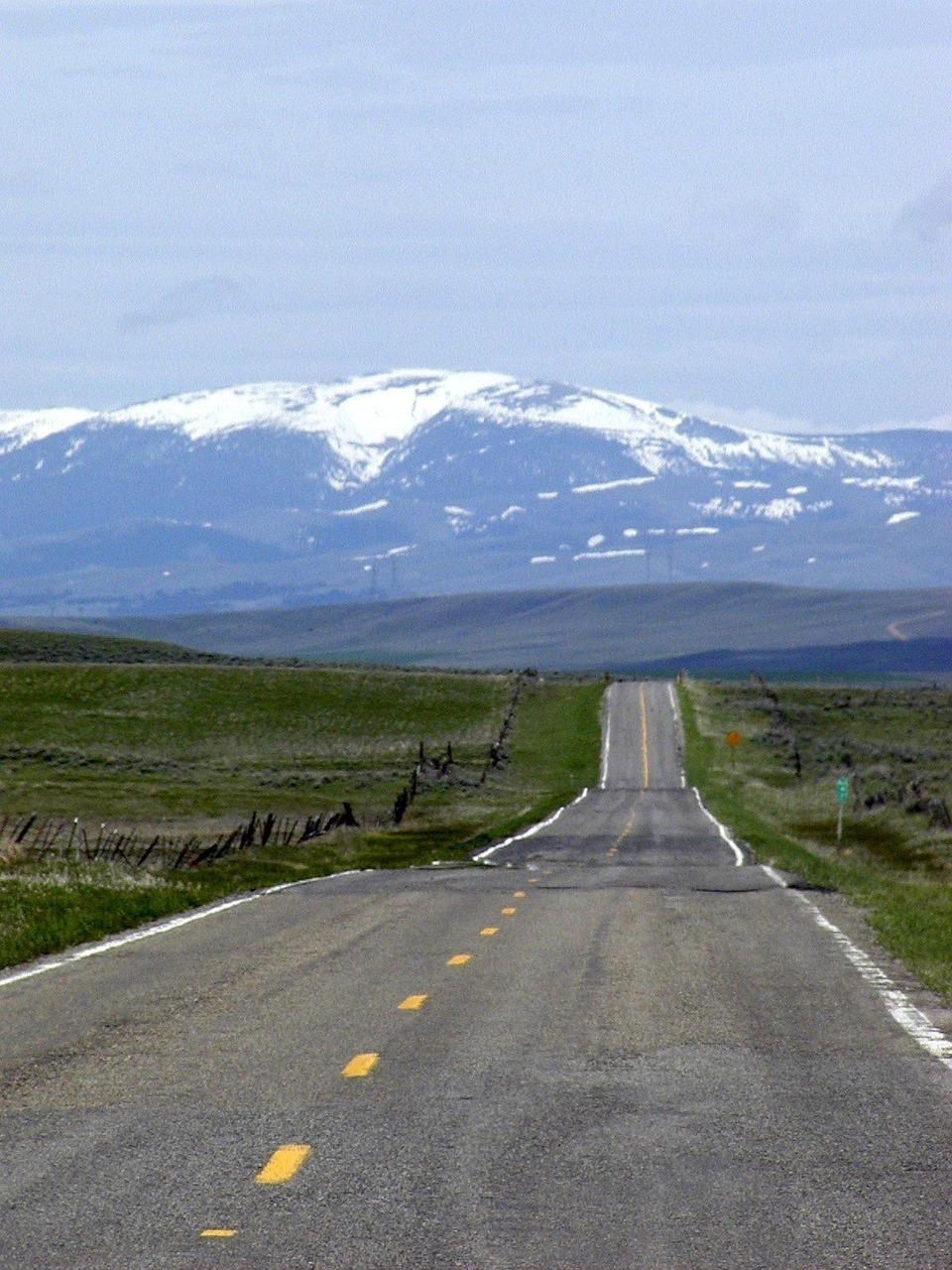 A long, open highway stretches through Montana's vast plains toward snow-capped mountains, capturing the state's rugged and scenic beauty.