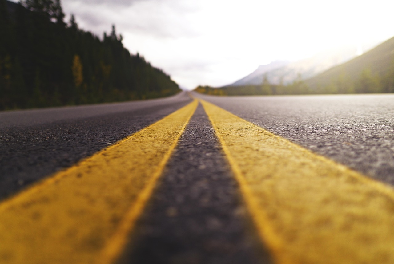 A low-angle view of a Montana highway captures the bold yellow centerlines stretching toward distant mountains, framed by dense pine forests and glowing in the golden light of dusk or dawn.